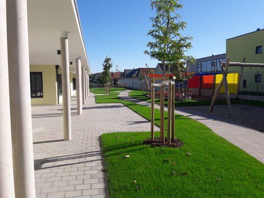 Ein sauberer, gepflegter Innenhof mit gepflasterten Wegen, grünem Rasen und Bäumen in der Mitte. Im Hintergrund sind ein gelbes Gebäude und eine rot, gelb und blaue Struktur zu sehen.