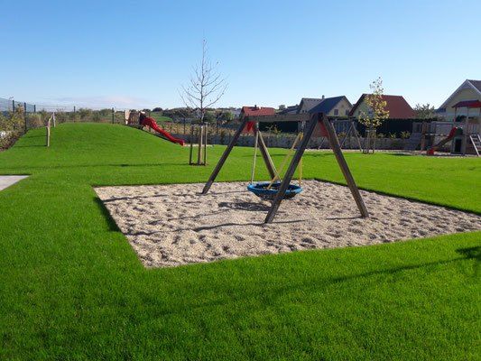 Ein Spielplatz mit grünem Rasen, Schaukeln und Rutschen, umgeben von Häusern und Bäumen an einem sonnigen Tag.