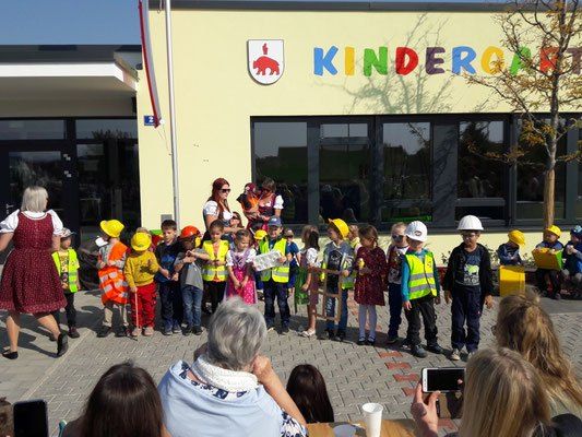 Kinder in Uniformen und Mützen stehen vor einem Kindergarten, Erwachsene im Hintergrund.