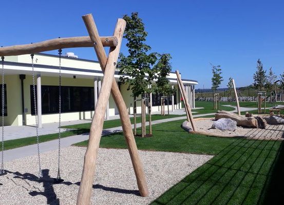 Ein Outdoor-Spielplatz mit Schaukeln und Sandkasten, umgeben von einem Gebäude mit Glasfenstern und Bäumen an einem sonnigen Tag.