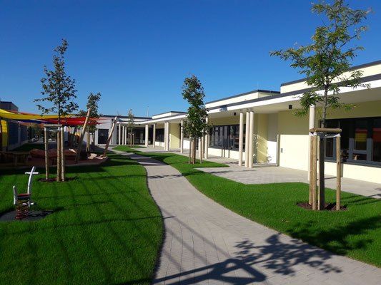 Ein Außenblick auf eine Schule mit einem langen Gehweg, grünem Gras und Bäumen. Der Weg ist gesäumt von jungen Bäumen und Sträuchern. Das Schulgebäude hat Glasfenster und Säulen.