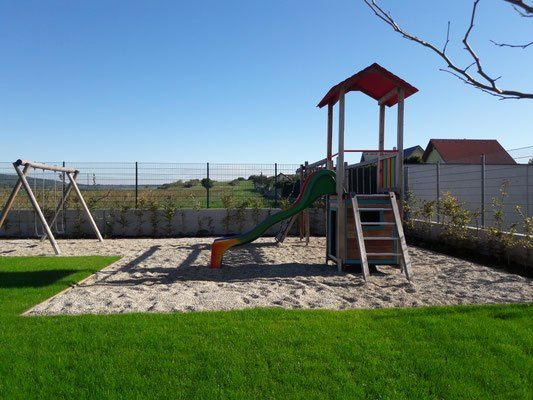 Ein Kinderspielplatz mit einer Rutsche, einer Schaukel und einem kleinen Klettergerüst, umgeben von Gras und einem Zaun.