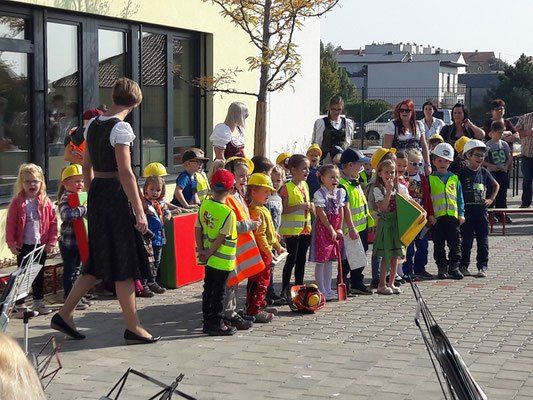 Eine Reihe von Kindern in Bauwesten, angeführt von einem Erwachsenen, steht vor einem Gebäude mit großen Fenstern. Einige halten Flaggen, und einige Erwachsene beobachten in der Nähe.