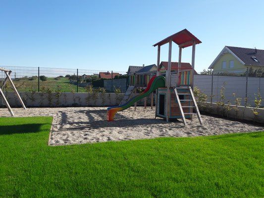 Ein Spielbereich für Kinder mit einer Rutsche und einem Klettergerüst, umgeben von einem Zaun, mit grünem Gras und Häusern im Hintergrund.