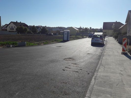 Eine ruhige Straße mit einem geparkten Auto, einer mobilen Toilette und Häusern im Hintergrund. Die Straße hat einen Bürgersteig mit einem dreieckigen Schild und einigen Fußabdrücken.