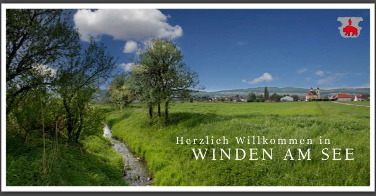 Eine ländliche Landschaft mit einem Bach, Bäumen und Hügeln unter einem blauen Himmel mit Wolken. Der Text 'Herzlich Willkommen WINDERN AM' ist prominent angezeigt.