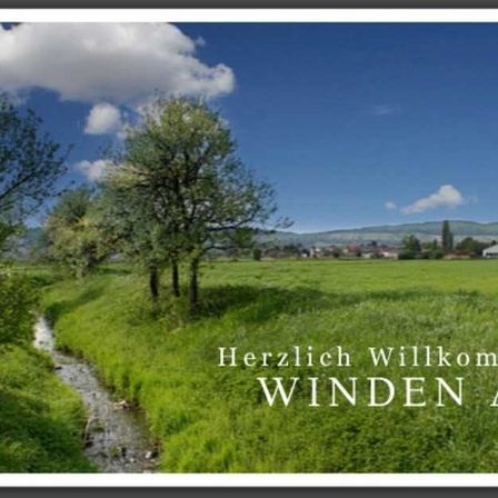 Eine ländliche Landschaft mit einem Bach, Bäumen und Hügeln unter einem blauen Himmel mit Wolken. Der Text 'Herzlich Willkommen WINDERN AM' ist prominent angezeigt.