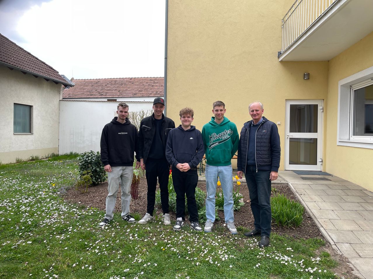 Fünf Männer stehen vor einem gelben Gebäude, lächeln und posieren für ein Foto in einem Garten.
