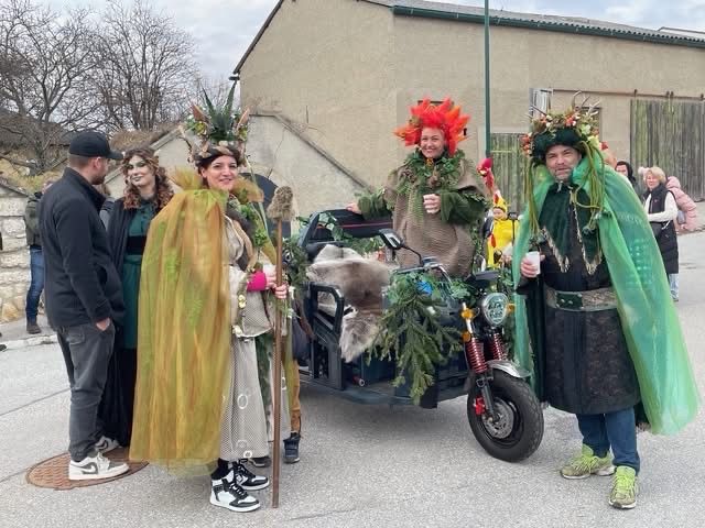 Eine Gruppe von Menschen in Kostümen steht vor einem Gebäude. Zwei tragen kräutige Kopfbedeckungen, einer hält einen Stab und ein anderer sitzt auf einem Motorrad.