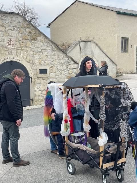 Ein Mann mit Rucksack steht neben einem Kinderwagen mit einem Regenschirm, und eine Frau in Kostüm sitzt im Wagen. Dahinter klettert eine Frau die Treppe hinauf, und ein Gebäude mit Steinmauer ist sichtbar.