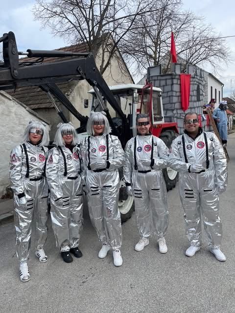 Fünf Menschen in silbernen Astronautenanzügen posieren vor einem Traktor und einer burgartigen Struktur. Ein Mann in einem blauen Overall steht in der Nähe, und eine Fahne ist an der Burg befestigt.