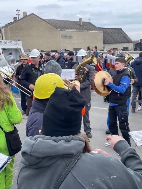 Eine Band spielt auf der Straße. Einige Leute tragen Hüte und halten Musikinstrumente. Ein Banner steht am Straßenrand.