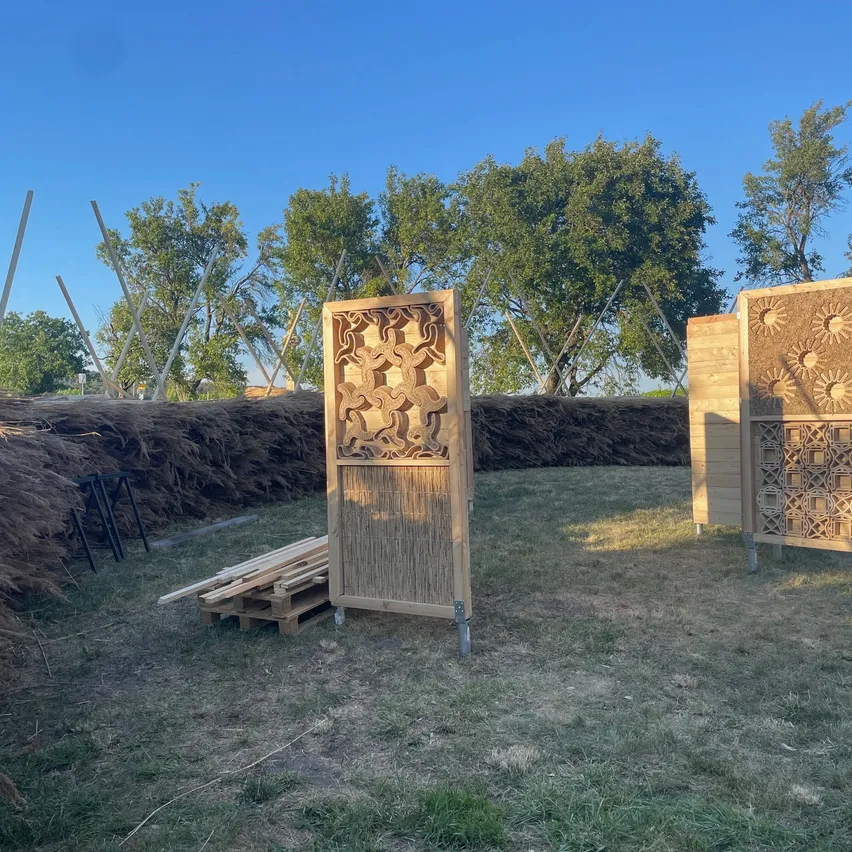 Zwei kunstvoll geschnitzte Holzplatten stehen in einer Außenumgebung mit einer Kulisse aus Bäumen und einem klaren blauen Himmel. In der Nähe befindet sich ein Holzstapel.