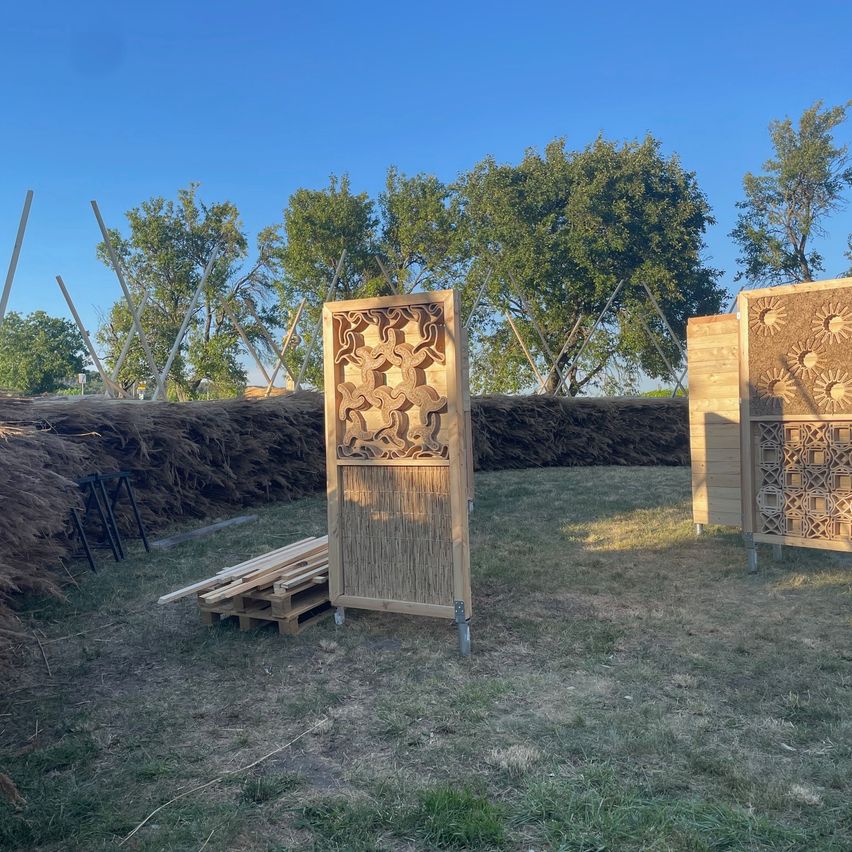 Zwei kunstvoll geschnitzte Holzplatten stehen in einer Außenumgebung mit einer Kulisse aus Bäumen und einem klaren blauen Himmel. In der Nähe befindet sich ein Holzstapel.