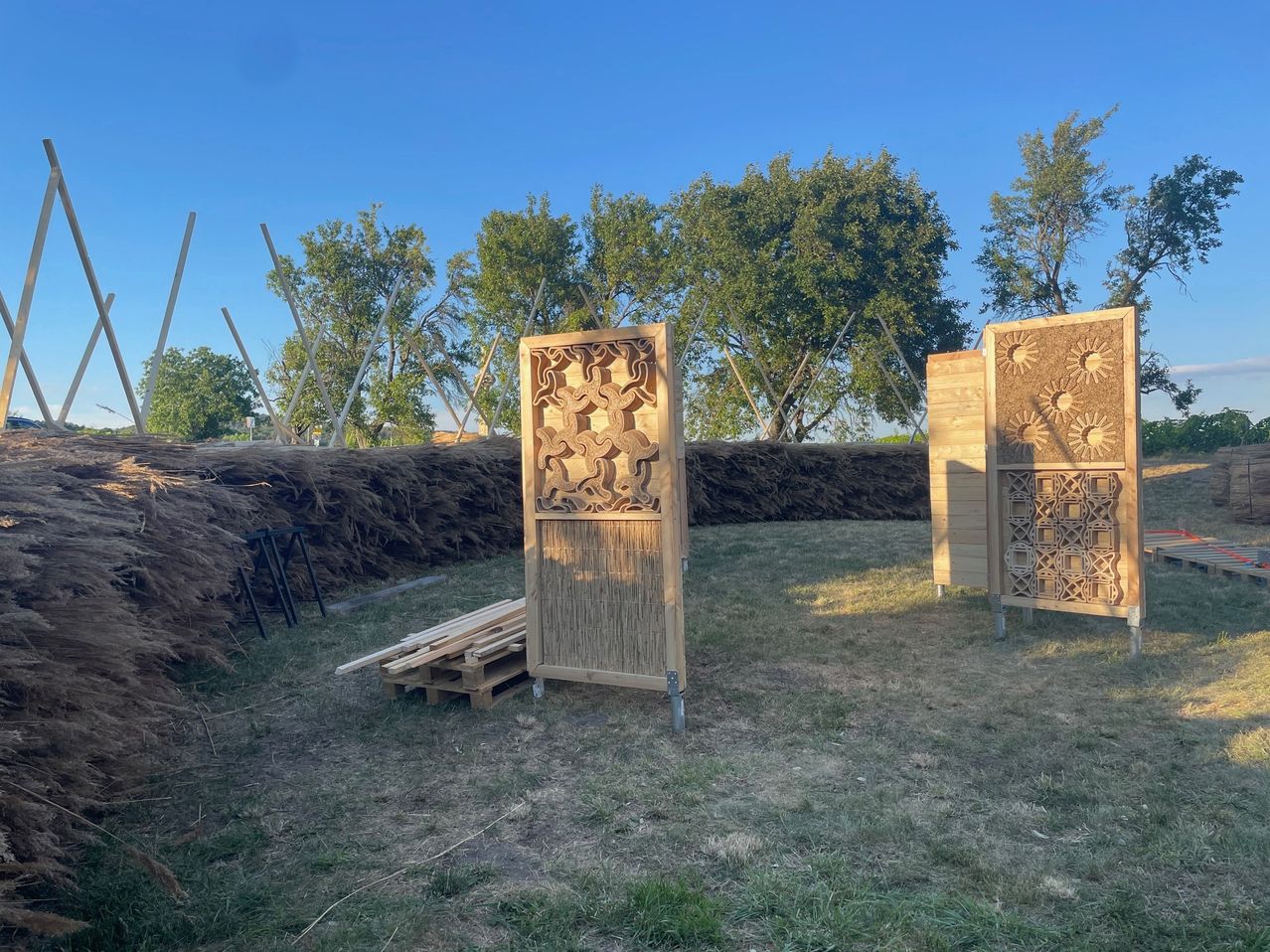 Zwei kunstvoll geschnitzte Holzplatten stehen in einer Außenumgebung mit einer Kulisse aus Bäumen und einem klaren blauen Himmel. In der Nähe befindet sich ein Holzstapel.