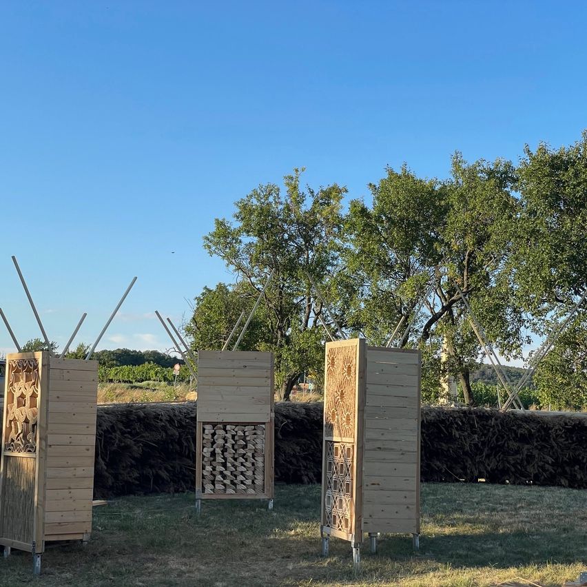 Drei Holzstrukturen mit geschnitzten Designs stehen auf einer Wiese mit einer Hecke und Bäumen im Hintergrund.