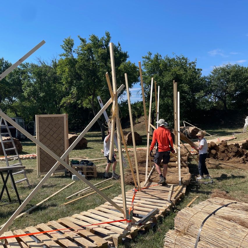 Eine Gruppe von Menschen baut eine Struktur aus Holzpfosten und Paletten auf einem Grasfeld. Bäume und ein klarer Himmel sind im Hintergrund zu sehen.