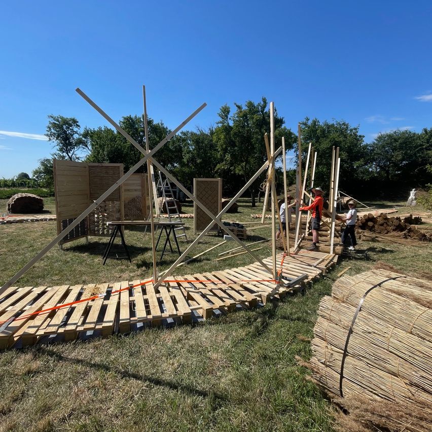 Drei Personen bauen einen Holzweg in einem Außenbereich mit Holzstrukturen und Bäumen im Hintergrund.