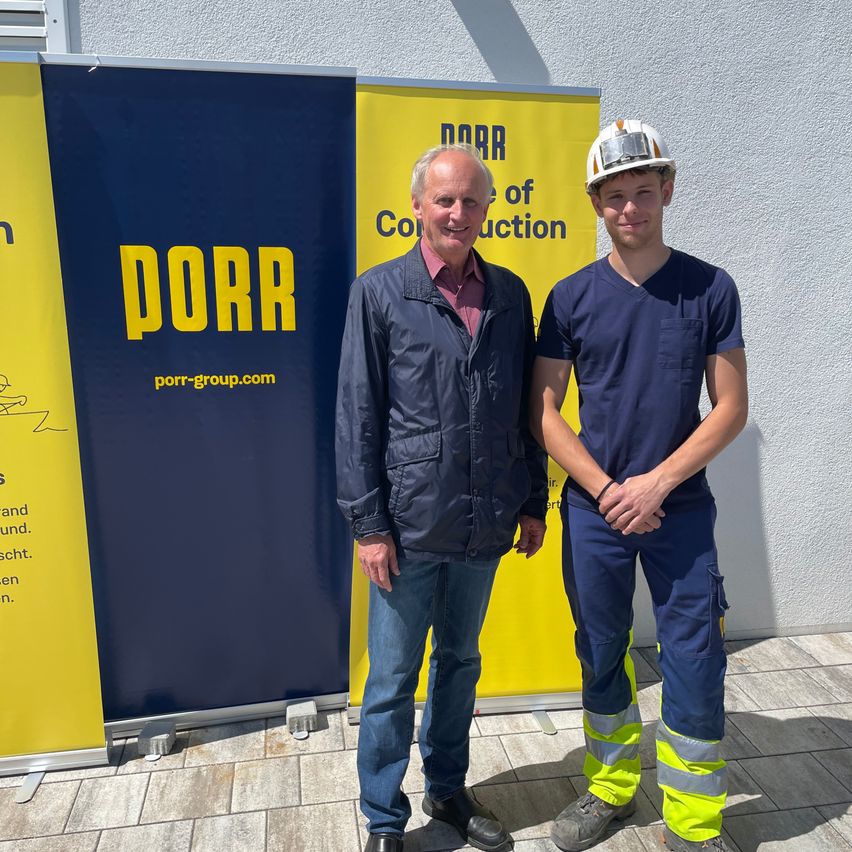 Two men stand next to each other in front of a PORR banner, wearing safety gear.