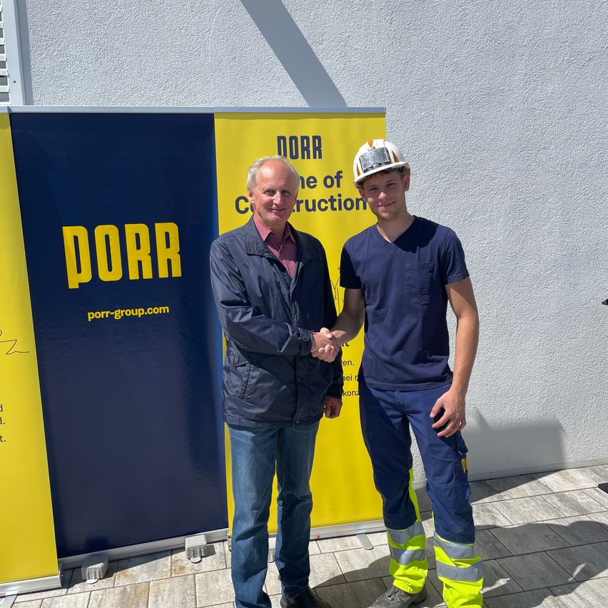 Two men stand in front of a PORR banner, one in a helmet and jacket, the other in a shirt and jeans, shaking hands.