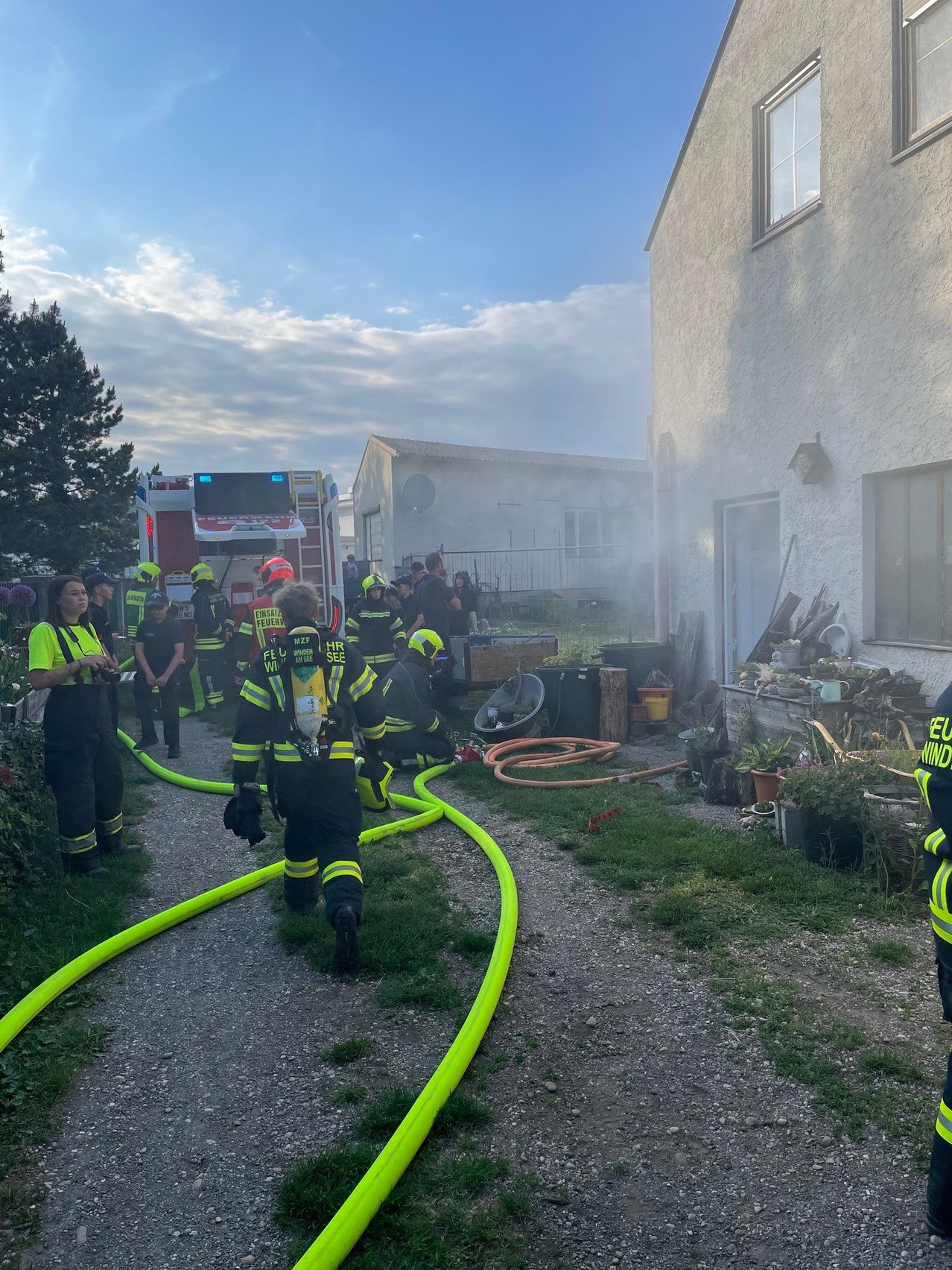 Firefighters in protective gear are working on a house fire. Smoke is coming from the building, and fire hoses are laid out on the ground.
