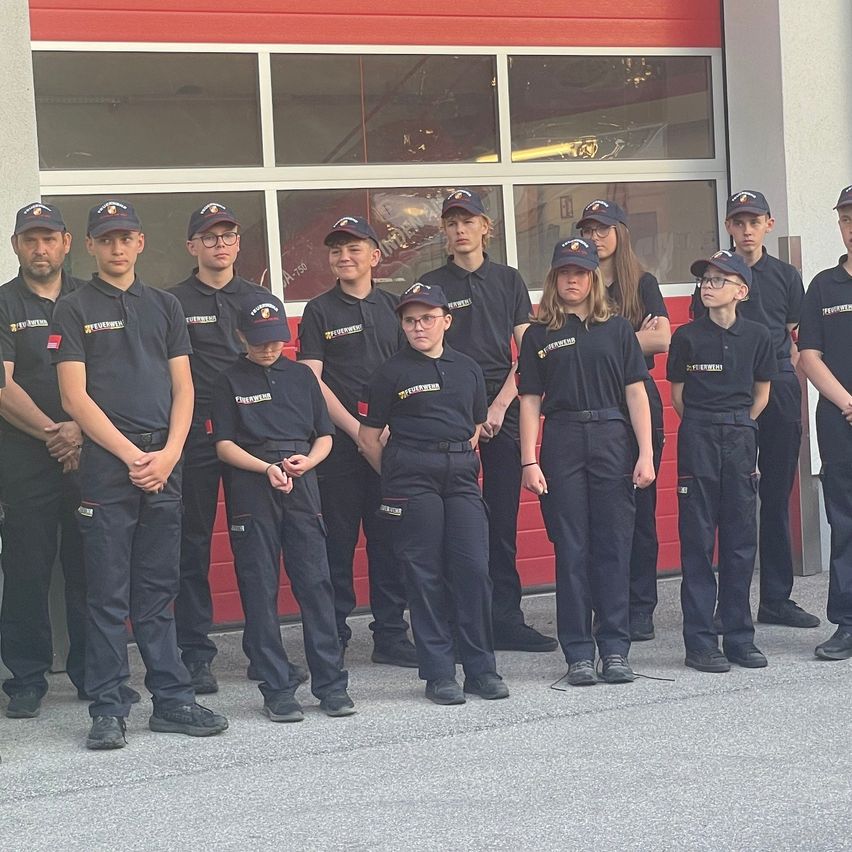 A group of young people in navy blue uniforms stand in front of a red fire station. They are wearing caps and some have glasses. A fire truck is visible in the background.