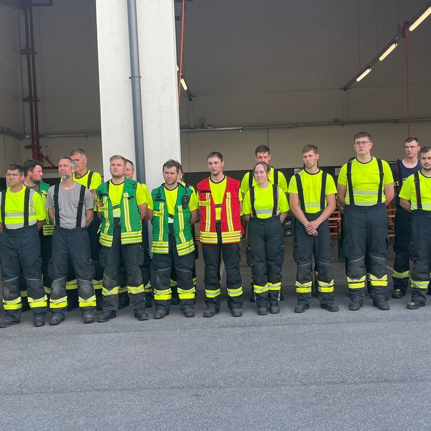 A group of firefighters, wearing safety gear, pose for a photo in front of a fire station.