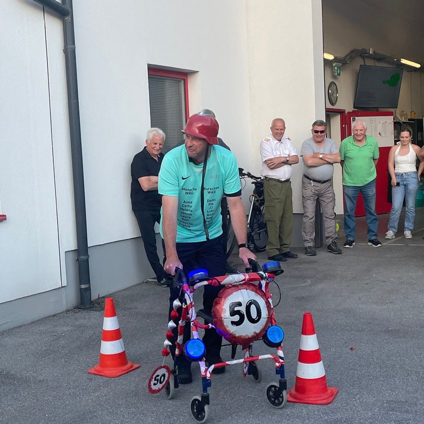 A man in a helmet is navigating a walker with a sign reading '50' between traffic cones, with onlookers watching.