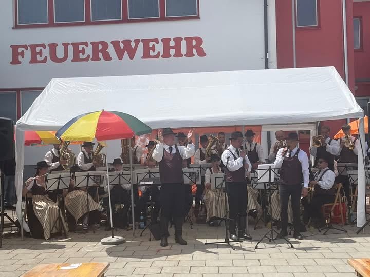 A brass band performs under a large tent on a paved outdoor area. The building behind them is labeled 'Feuerwehr'.