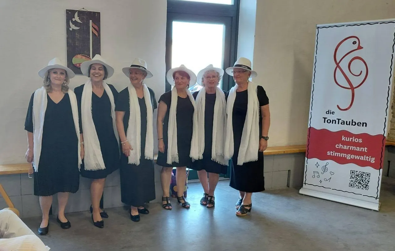 Fünf Frauen lächeln vor einer weißen Wand mit einer Tür und einer Bank. Sie tragen schwarze Kleider, weiße Schals und weiße Hüte.