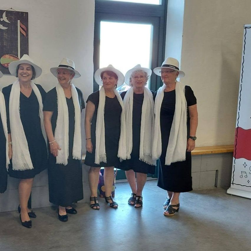 Fünf Frauen lächeln vor einer weißen Wand mit einer Tür und einer Bank. Sie tragen schwarze Kleider, weiße Schals und weiße Hüte.