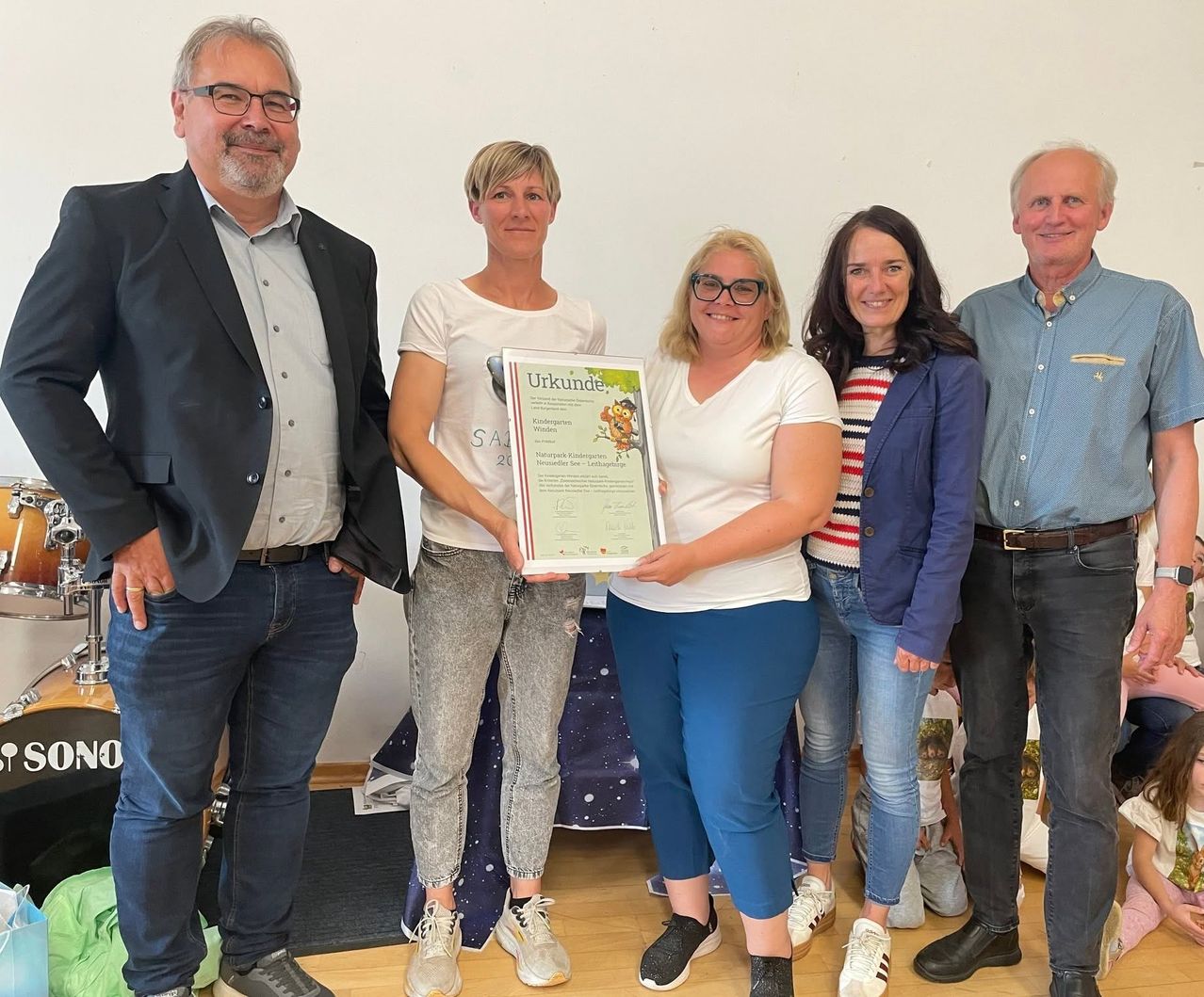 Fünf Personen stehen zusammen für ein Foto mit einer Frau, die einen gerahmten Preis hält. Die Frau rechts lächelt, während der Mann links nach unten schaut. Hinter ihnen hat eine weiße Wand ein farbenfrohes Design.