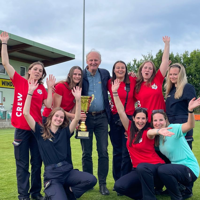 A group of women and a man, dressed in red and black uniforms, are posing for a picture on a field. The man is holding a trophy.