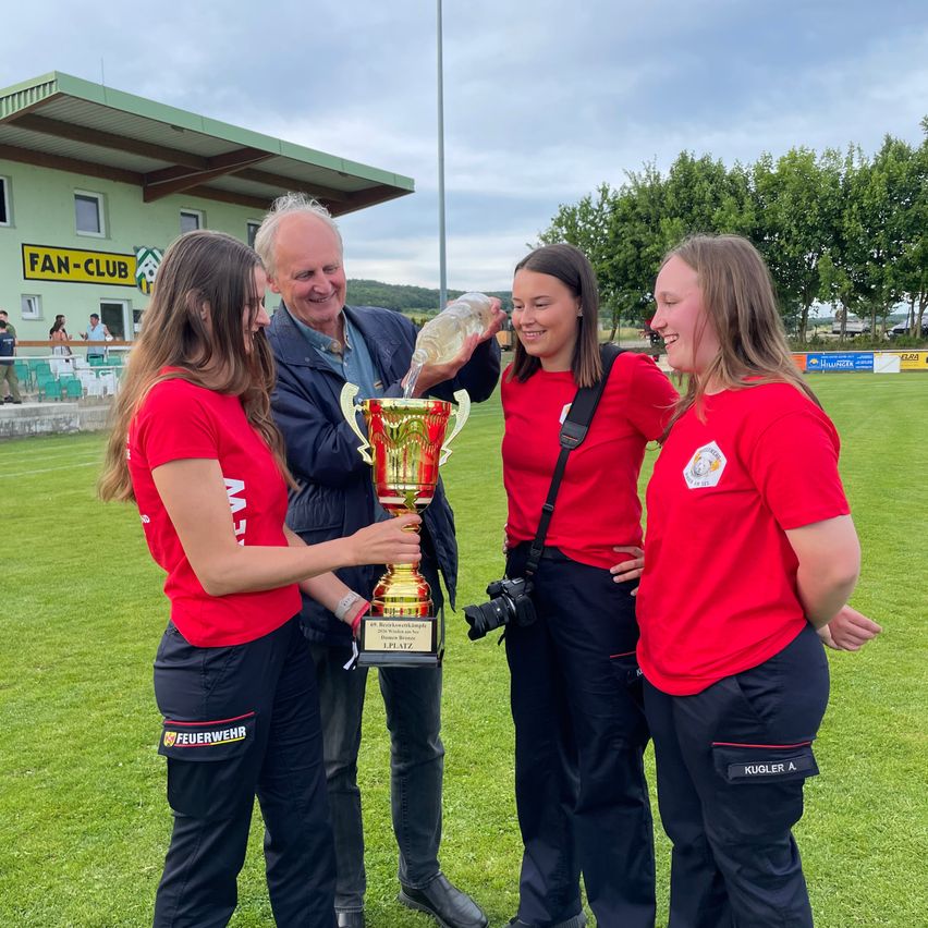 Vier Personen in roten T-Shirts stehen auf einem Feld und halten eine Trophäe. Ein Mann gießt Wasser über die Trophäe. Hinter ihnen steht ein Gebäude mit einem Fanclub-Schild. Bäume und ein Zaun sind im Hintergrund.