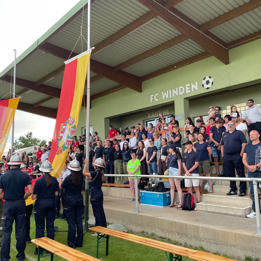 Eine Gruppe von Menschen, wahrscheinlich Fans, steht auf den Stufen eines Stadions. Sie tragen Uniformen, Mützen und Schuhe. Einige halten Fahnen, während andere das Spiel verfolgen.