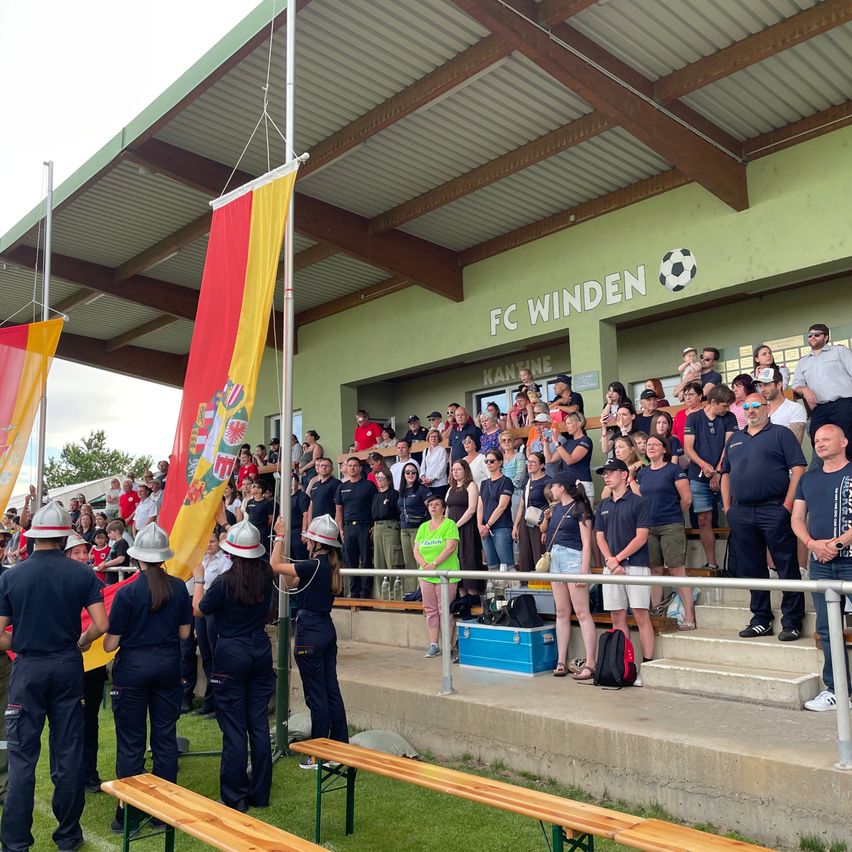 Eine Gruppe von Menschen, wahrscheinlich Fans, steht auf den Stufen eines Stadions. Sie tragen Uniformen, Mützen und Schuhe. Einige halten Fahnen, während andere das Spiel verfolgen.