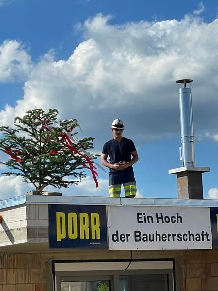 Ein Mann steht auf einem Dach, trägt einen Helm und steht vor einem Schild mit der Aufschrift PORR unter einem teilweise bewölkten Himmel.