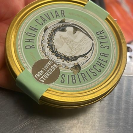 Eine Hand hält eine Dose Rhön Kaviar mit Fischdesign. Das Etikett zeigt 'Von lebenden Sturgeonen' und 'Sibirischer Stör'. Die Dose hat einen goldenen Rand.