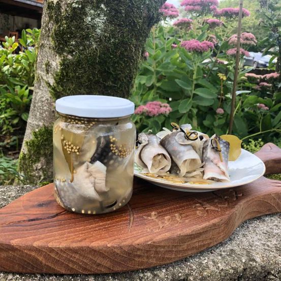 Ein Glas mit eingelegten Fischen und ein Teller mit Fischrollen stehen auf einem Holzbrett draußen neben einem Baum und Blumen.
