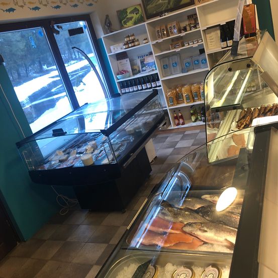 Ein Indoor-Fischmarkt mit Glastheken, die Fische präsentieren, begleitet von Regalen, die mit verschiedenen Artikeln bestückt sind, und einem Fenster, das eine verschneite Landschaft zeigt.