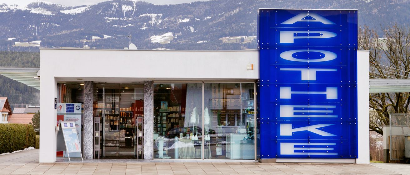 Eine Apotheke mit einem blauen Schild und Glastüren steht vor verschneiten Bergen.
