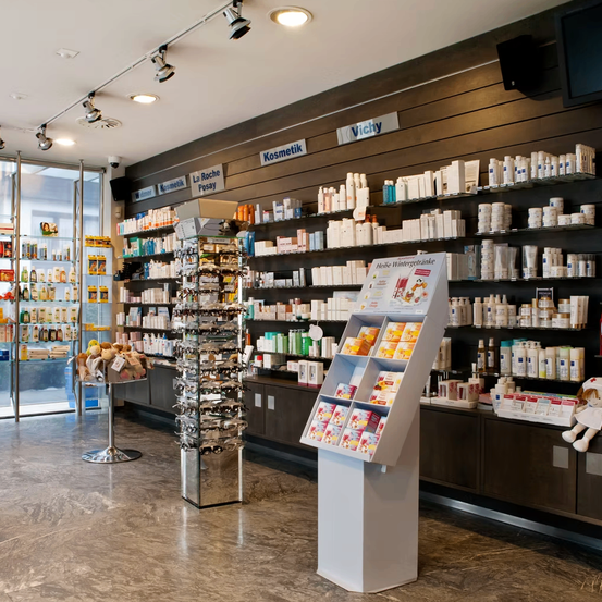 Ein Apotheken-Innenraum mit Regalen, die mit verschiedenen Hautpflege- und Kosmetikprodukten bestückt sind. Die Regale haben Etiketten wie 'Vichy' und 'Kosmetik'. Eine Anzeige mit mehreren Produkten steht in der Mitte und ein Plüschtier ist auf der linken Seite.