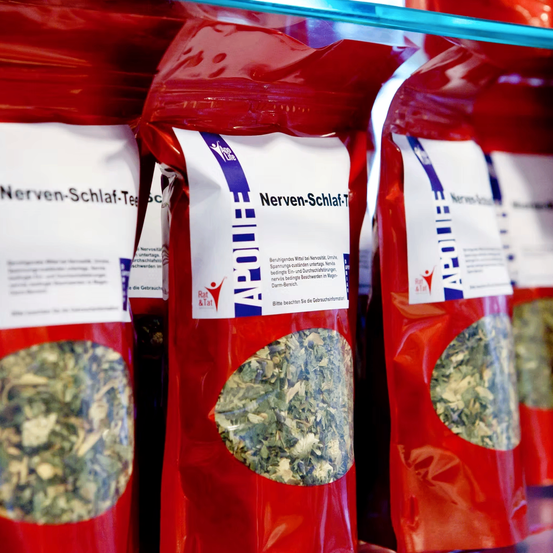 Mehrere rote Kräutertee-Pakete sind auf einem Regal aufgereiht. Das mittlere Paket ist mit 'Nerven-Schlaf-Tee' beschriftet und enthält getrocknete Kräuter.