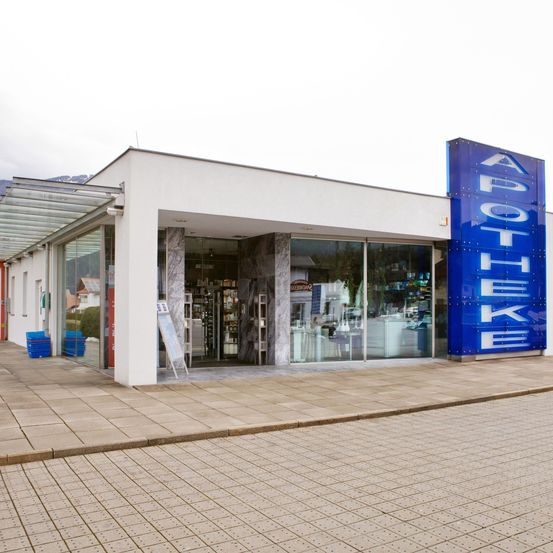 Außenansicht einer Apotheke mit einem blauen Schild, das 'APOTHEKE' steht. Die Apotheke hat Glaswände, und im Inneren befinden sich Gegenstände auf Regalen.