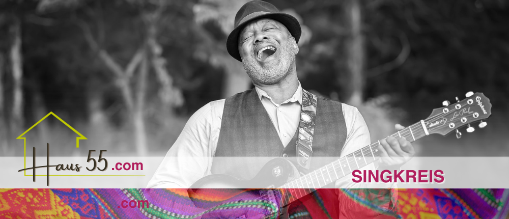 Ein Schwarz-Weiß-Foto eines Mannes, der eine Gitarre spielt und singt, während er die Augen geschlossen hat. Er trägt einen Hut, eine Weste und hat einen bunten Hintergrund vor sich. Der Text lautet 'Singkreis'.