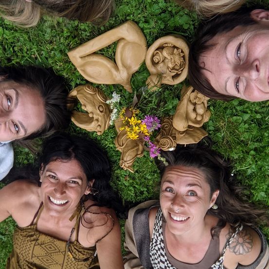 Fünf Frauen liegen lächelnd auf Gras mit Tonfiguren von Tieren und Pflanzen. Die Frau in der Mitte hat eine Blumenkette.