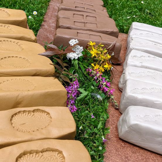 Eine Reihe von Lehmziegeln mit verschiedenen Designs ist auf einem Grasbereich ausgestellt. Die Ziegel sind in einem Zickzackmuster angeordnet.