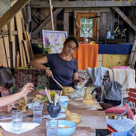 Zwei Frauen in einem rustikalen Studio, eine malt eine Tonfigur, während die andere an einer Skulptur arbeitet. Verschiedene Kunstmaterialien sind auf dem Tisch und den Regalen.