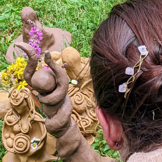 Eine Frau hält eine Tonfigur eines Babys auf dem Schoß. Mehrere Tonfiguren sind um sie herum, einschließlich einer Figur mit Blumen. Sie schaut in die Kamera.