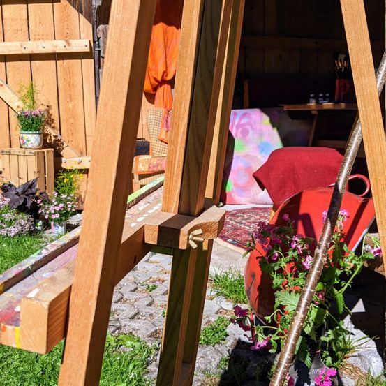 Ein Holzstaffelei steht in einem Garten mit bunten Blumen. Dahinter ist ein Holzschuppen sichtbar, in dem ein Gemälde auf dem Boden liegt. Ein roter Stuhl und ein Tisch mit Flaschen sind drinnen.